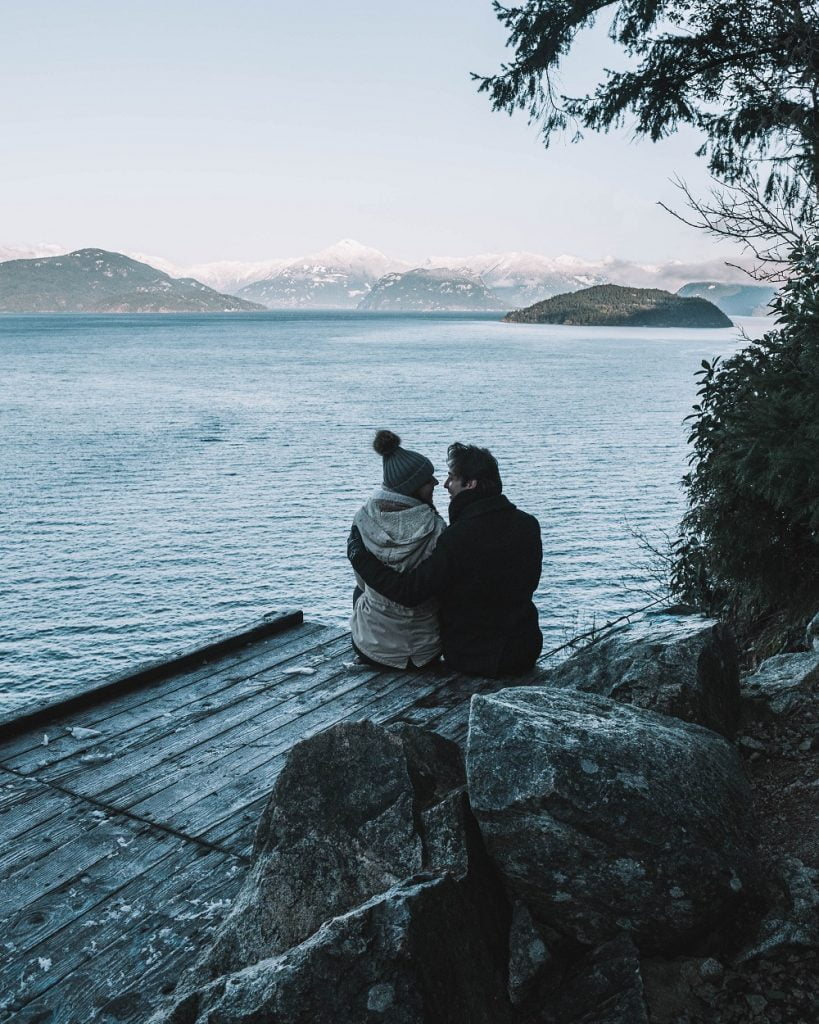 couple sitting on jeti looking out over lake with snow covered mountains in distance