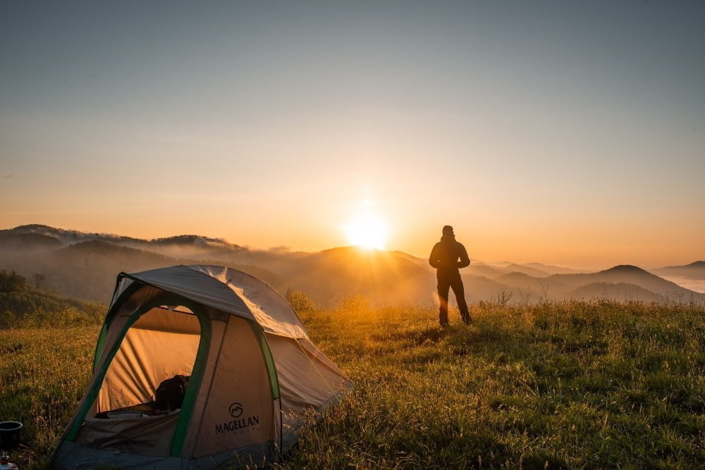 Sustainable Outdoor Adventure: person in travel geaer watching the sun rise over the mountains by their tent