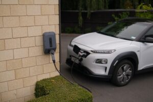 Impact of Electric Vehicles: white electric vehicle plugged in by sandstone brick wall outside a house