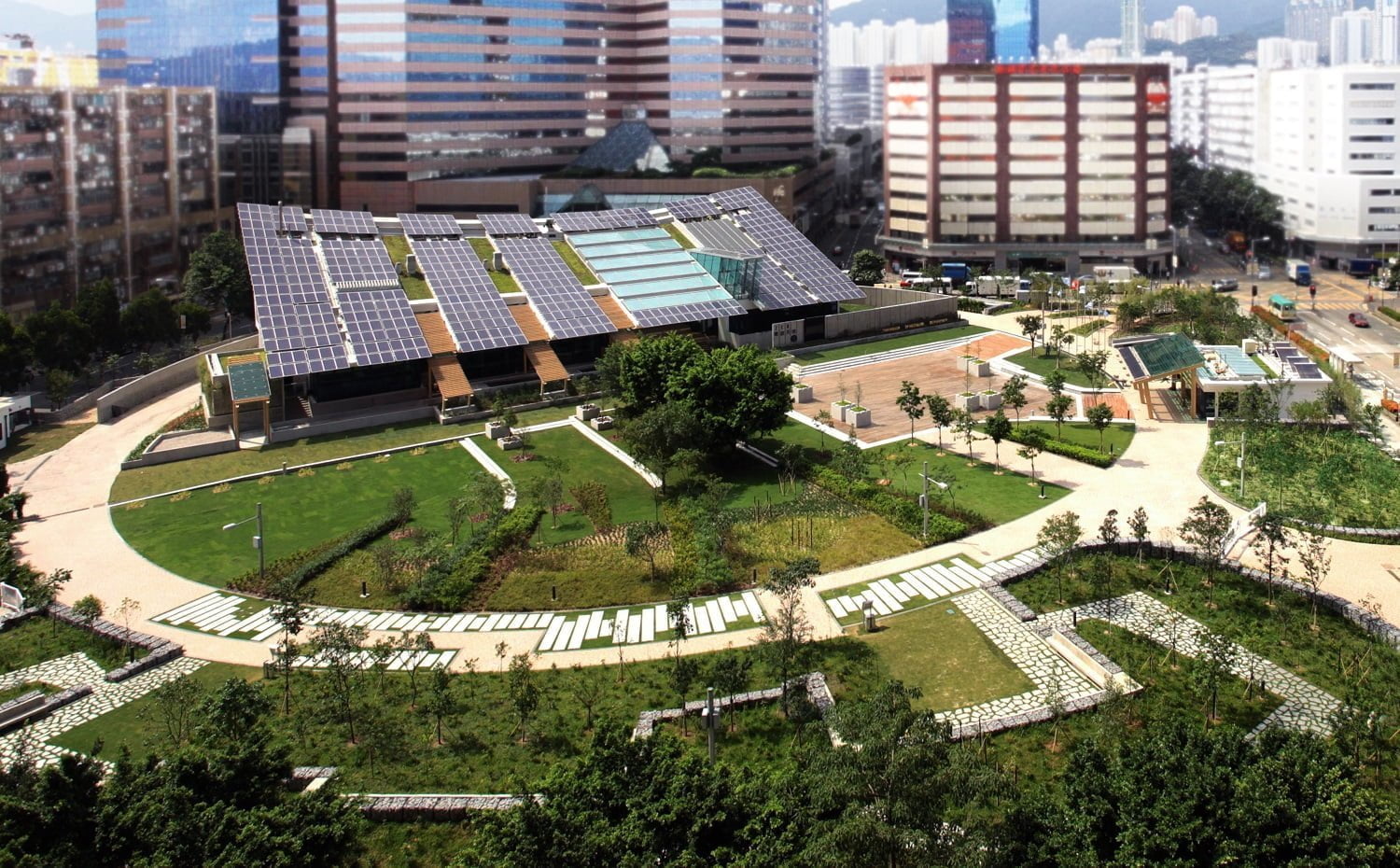 A wide roof covered with solar panels, with greenery around and buildings beyond